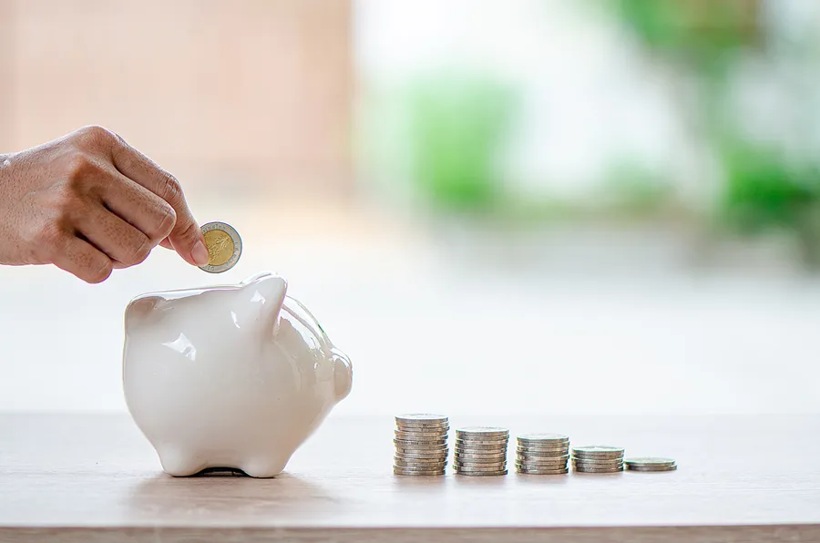 Mano echando una moneda en una hucha, junto a varias pilas de monedas ordenadas